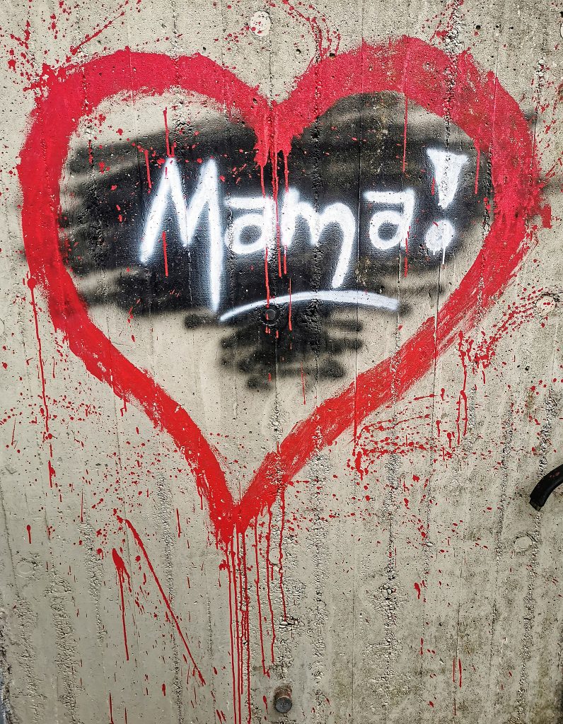Graffito: Ein Herz mit rotem Rand auf einer kahlen Mauer. Die rote Farbe verläuft nach unten, Darin eine schwarze unregelmäßige Fläche. Mit weißer Schrift darauf der Schriftzug Mama! weiß unterstrichen.