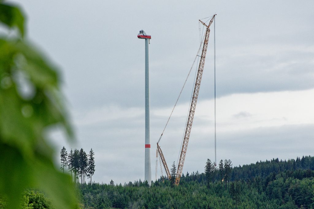 Im Vordergrund links unscharf Blätter eines Baums in der Bildmitte der Turm einer Windkraftanlage mit dem Fuß im Wald samt riesigem Kran rechts vom Turm. Die Flügel der WKA fehlen noch.