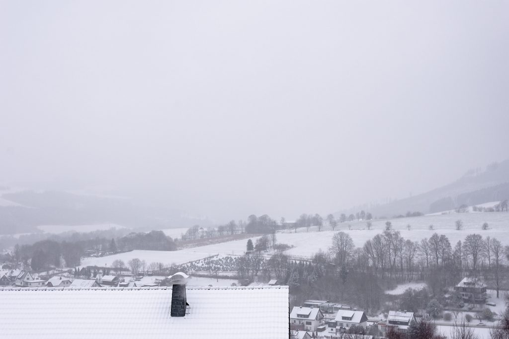 Blick auf eine verschneite Hügellandschaft mit Baumbeständen. Im Vordergrund das mit Schnee bedeckte Dach eines Hauses plus Schornstein.