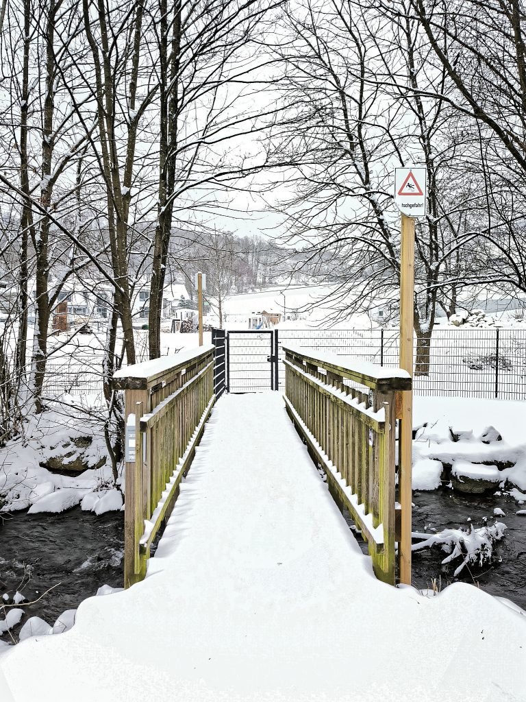 Eine hölzerne Fußgängerbrücke führt über einen Bach. Im Hintergrund ein Zaun; eine verschneite, hügelige Landschaft in der Ferne. Grauer Himmel. Rechts vor an der Brücke ein Holzpfahl mit einem kleinen Warnschild: Rutschgefahr!