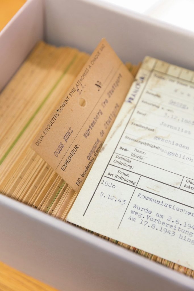 Die geöffnete weiß-graue Schublade eines Archivschranks. Darin sind hintereinander viele  braunorangene Karteikarten mit getippten Angaben. Eine weißliche Karteikarte liegt zur Veranschaulichung vornan horizontal über den senkrechten anderen Karten. Man kann noch einzelne Wörter wie "kommunistischer" lesen.