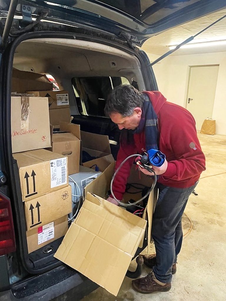 Ein Mann mit rotem Kapuzenpullover ist über einen Pappkarton gebeugt, in welchem sich verschiedene medizinische (?) Hilfsmittel befinden. Die rechte Hand greift in den Karton. die linke hält einen Schlauch und eine Blutdruckmessgerät (?) Der Kofferraum des Caddys ist voller Kisten. Es ist kaum noch Platz. Die Heckklappe ist nach oben geöffnet. Der beschriebene Karton steht auf dem Ende des Kofferraumbodens. Das linke Knie des Mannes klemmt ihn zur Stabilisierung ein.