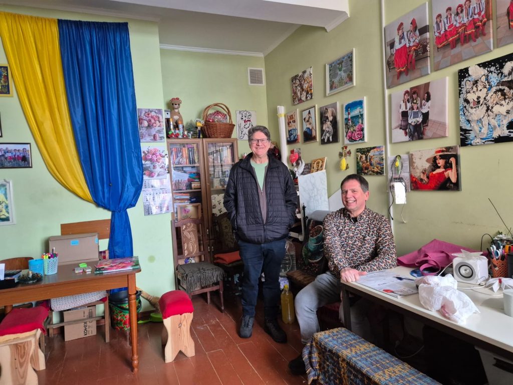 Zwei Männer in einem bunt gestalteten Raum. Viele Bilder an der Wand. Links ein Vorhang in den Farben der Ukraine (gelb-blau).