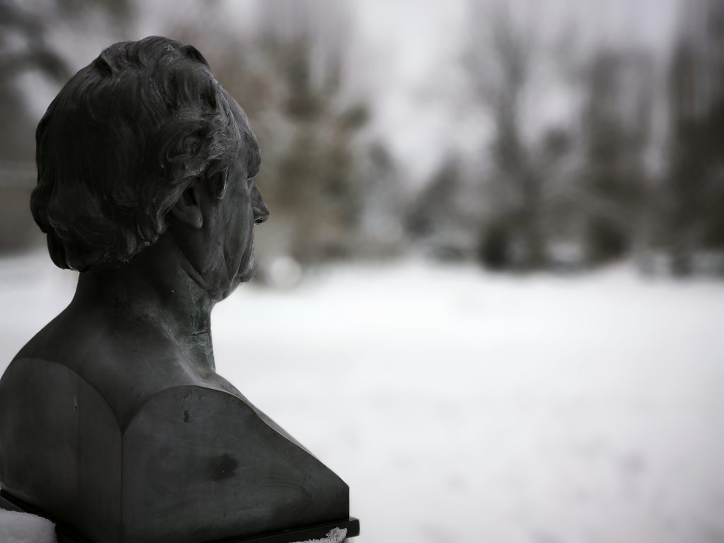 Ein Dichter-Büste von halb-rechts-hinten fotografiert. Der Blick ist in den unscharfen Hintergrund, eine Mischung aus Schnee und Sträuchern, gerichtet.