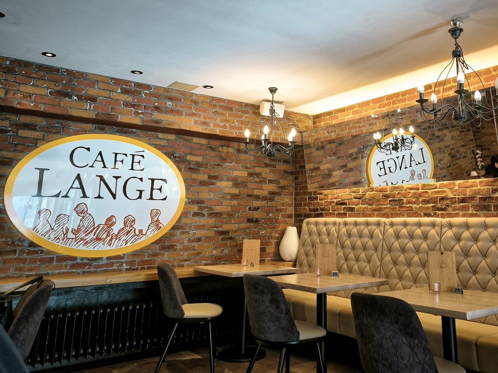 Innenraum des Cafés Lange. Der Name steht auf einem großen ovalen Schild, welches sich in einem Spiegel über den Sitzbänken rechts spiegelt.
Weiße Decke mit "Kronleuchtern" und geziegelte Wände.