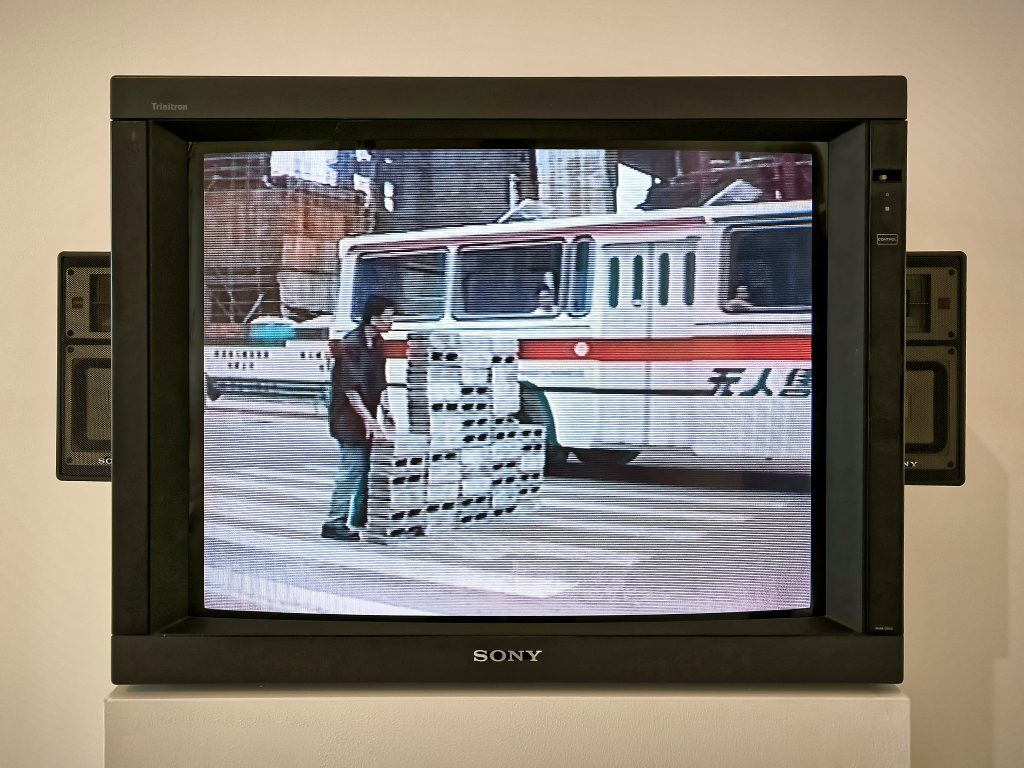 Ein Fernsehmonitor. Ein Mann baut eine Mauer quer zu einem Zebrastreifen hinten ab. Im Hintergrund ein Bus und dahinter werden Häuser gebaut.
