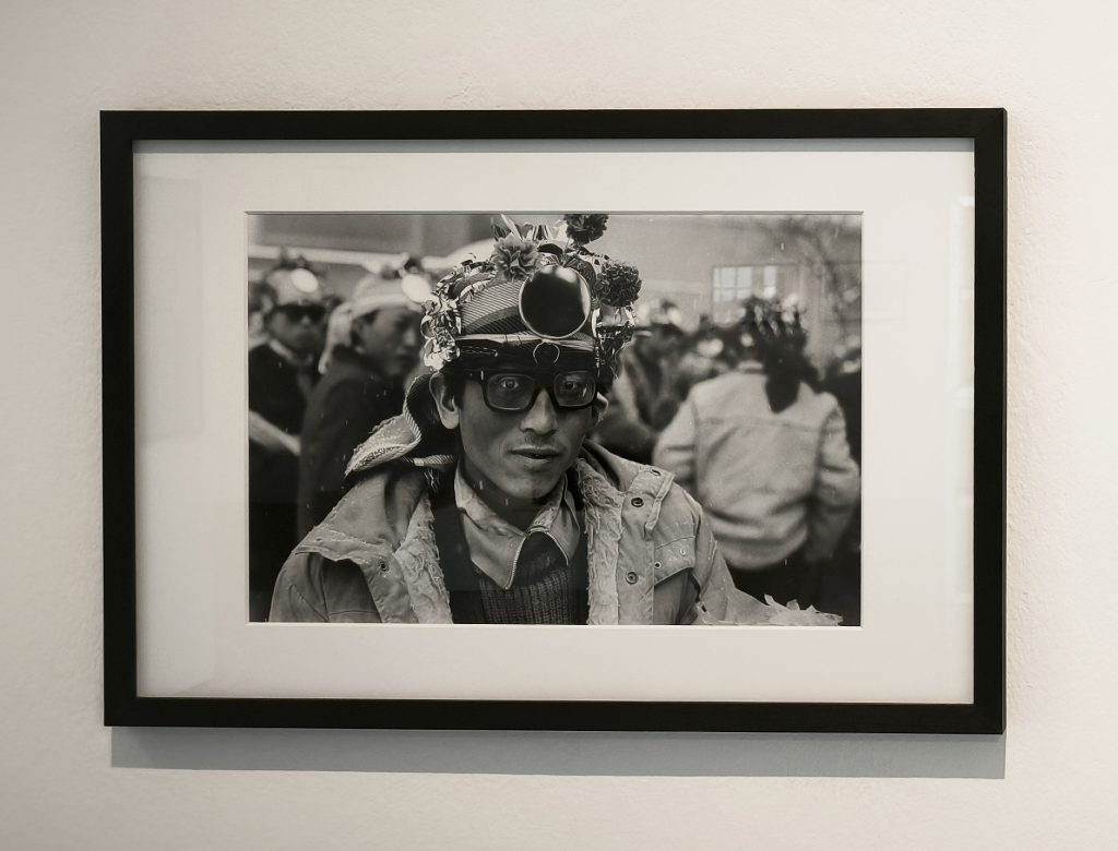 Ein Mann schaut von einer Fotografie zum Betrachter. Er hat eine Fantasiemütze/Hut auf. Im unscharfen Hintergrund stehen und sitzen wahrscheinlich feiernde Menschen, die ähnliche Hüte tragen.

Auffällig sind die großen, dicken Brillengläser der Mannes im Vordergrund.