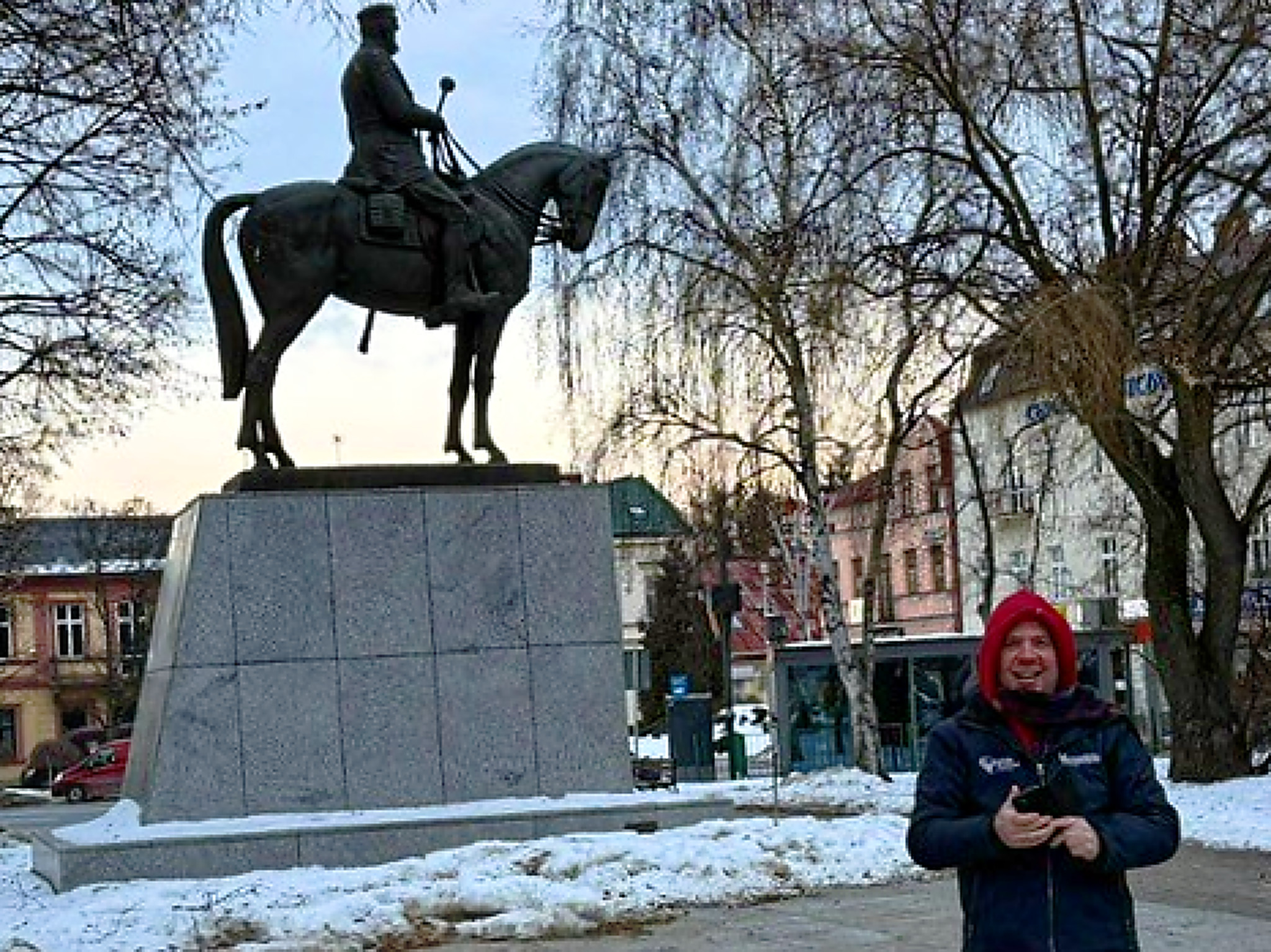 Ein Mann steht auf einem städtischen Platz. Eine rote Kapuze über dem Kopf, ein Handy in der Hand. Er lacht. Hinter ihm links ein riesiges Reiterdenkmal, grauer Sockel, Schwarzes Pferd und Reiter mit Marschallstab in der linken Hand.
