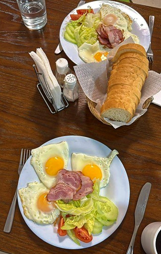Ein Tisch mit zwei großen weißen Tellern. Darauf jeweils drei Spiegeleier, drei Fleischscheiben, Tomaten, Gurken, Salat. Ein Korb mit geschnittenem Baguette, Kaffee, Wasser, Messer, Gabel
