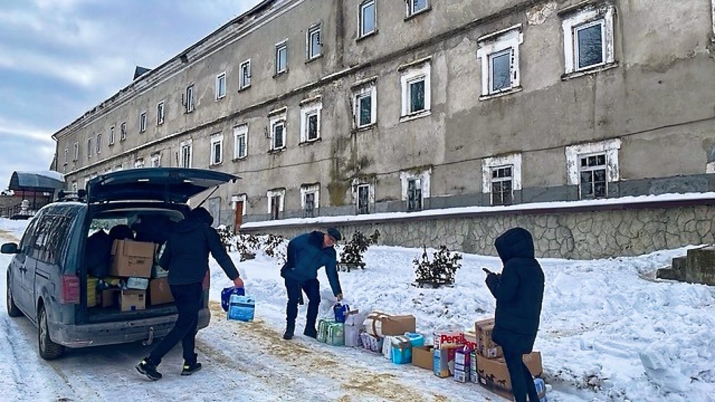 Ein großer grauer Klotz (Gebäude) rechts, davor die verschneite Straße mit dem Caddy, aus dem Hilfsgüter an den Rand getragen werden.
