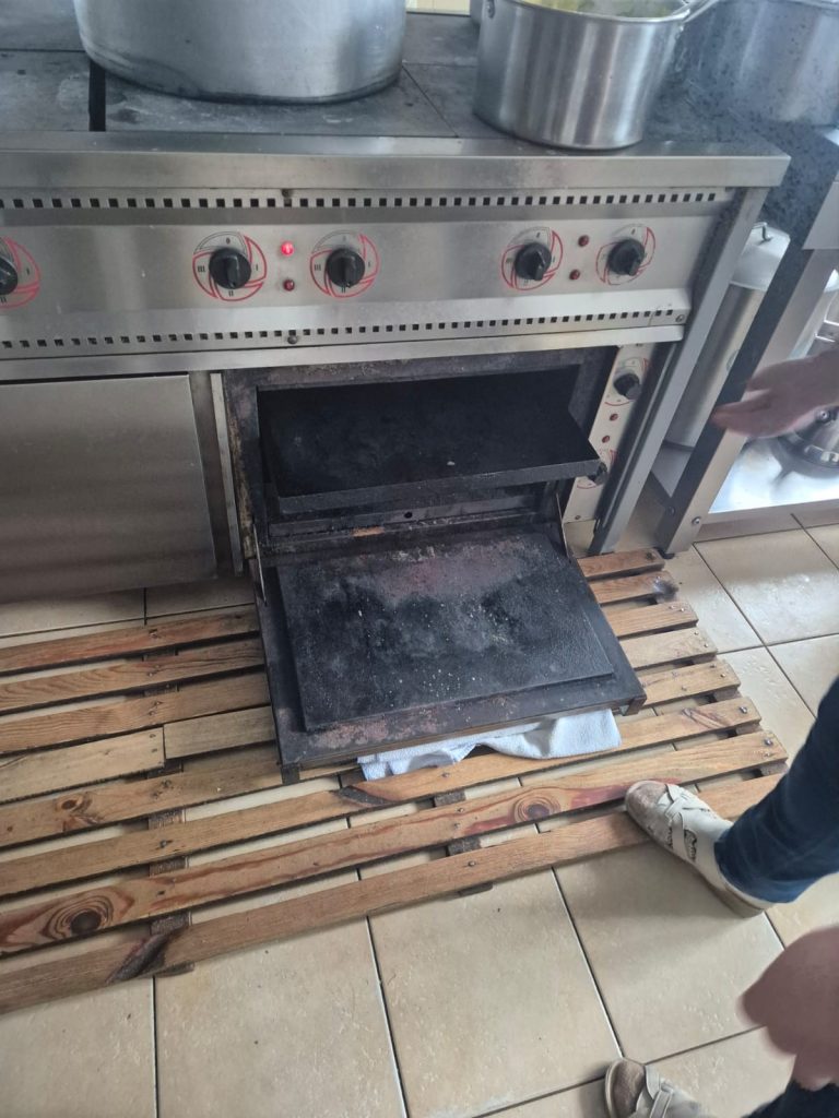 Die verrostete Klappe eines alten Herdes ist geöffnet. Davor ein Holzrost auf dem Boden.