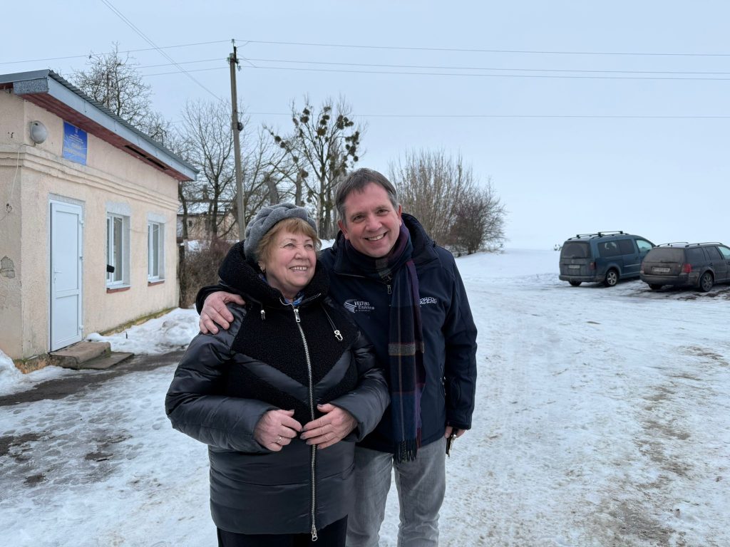 Ein Mann und eine Frau stehen neben einem niedrigen Gebäude im Schnee auf einem Parkplatz. Der Mann links hat seinen rechten Arm über die Schultern der etwas kleineren Frau gelegt. Im Hintergrund rechts sind zwei Autos zu sehen: Links ein Caddy, rechts ein Combi.