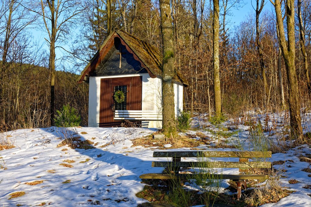 Eine kleine Kapelle liegt zwischen Bäumen im Wald. Direkt vor der Kapelle rechts ist eine weiße Bank. Der Schnee zwischen den Bäumen ist löchrig; brauen Flecken mit Wiese. GAnz vorn rechts steht noch eine braune Holzbank, mit Flechten überzogen.