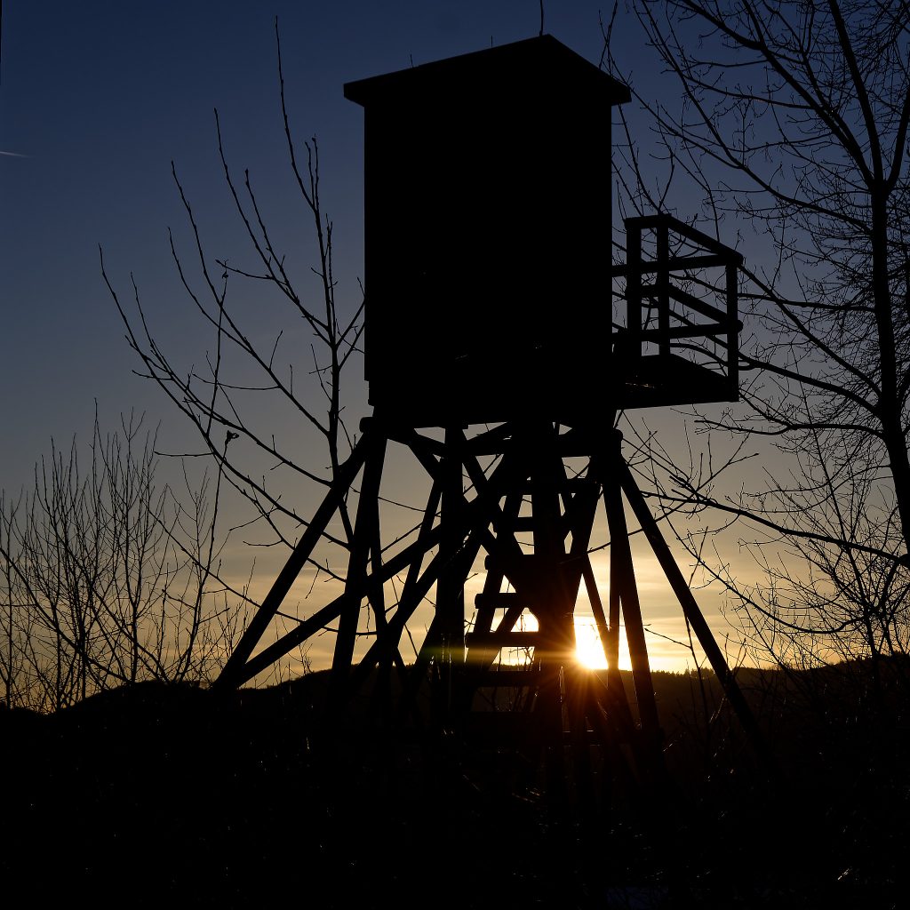 Eine Jagdkanzel ragt in den dunklen Abendhimmel. Am Fuß der Treppe ist noch ein Teil der untergehenden Sonne zu sehen.