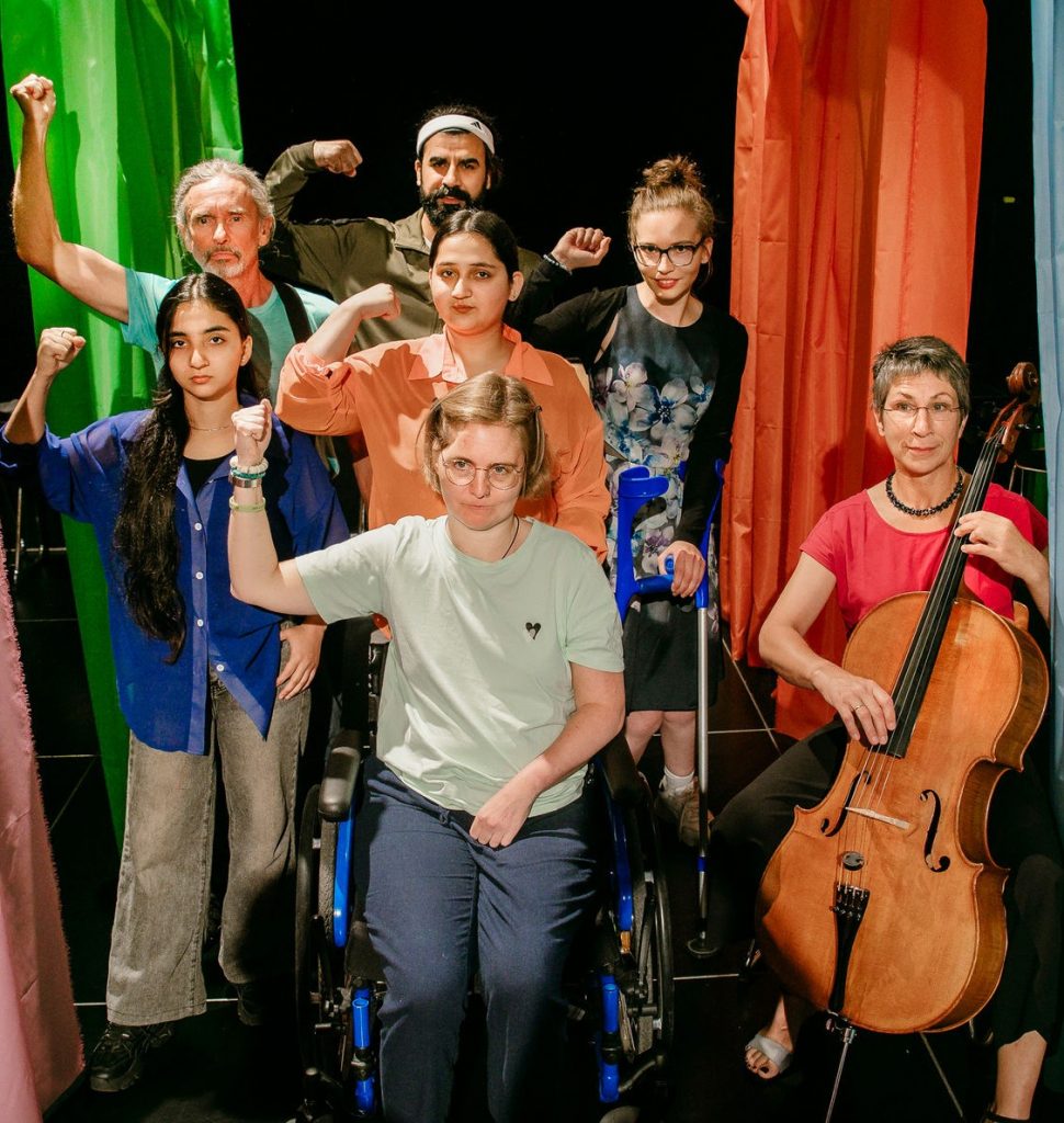 Auf einer Theaterbühne befinden sich sieben Personen. In der ersten Reihe der Gruppe sitzt mittig eine junge Frau im Rollstuhl, rechts sind eine weitere Frau mit Cello. In der zweiten und dritten Reihe sind weitere Personen zu sehen. Bis auf die Cello-Spielerin haben alle die Fäuste wie zum Kampf gereckt. Links hängt ein grüner rechts ein rot-orangener Vorhang auf die Bühne hinunter. Die Menschen wirken sehr entschlossen und strahlen trotzdem, auch wegen ihrer farbenfrohen Kleidung, Optimismus aus.