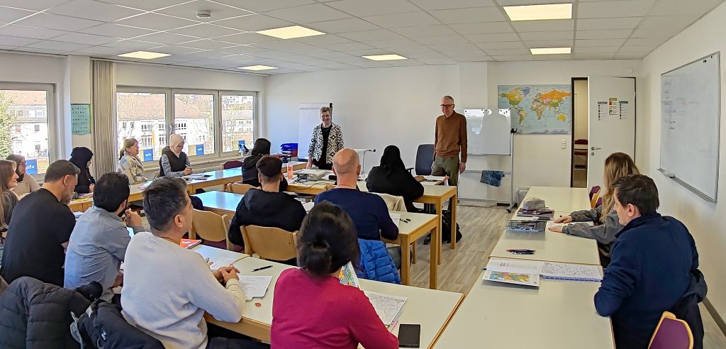Blick in einen hellen Unterrichtsraum mit weißen Wänden, weißer Decke und hellen Tischen. Teilnehmer*innen sitzen im U mit zwei Sprossen. Links ist eine Fensterseite, rechts eine weiße Tafel. Vorne (im Bildhintergrund) stehen zwei Dozent*innen.
