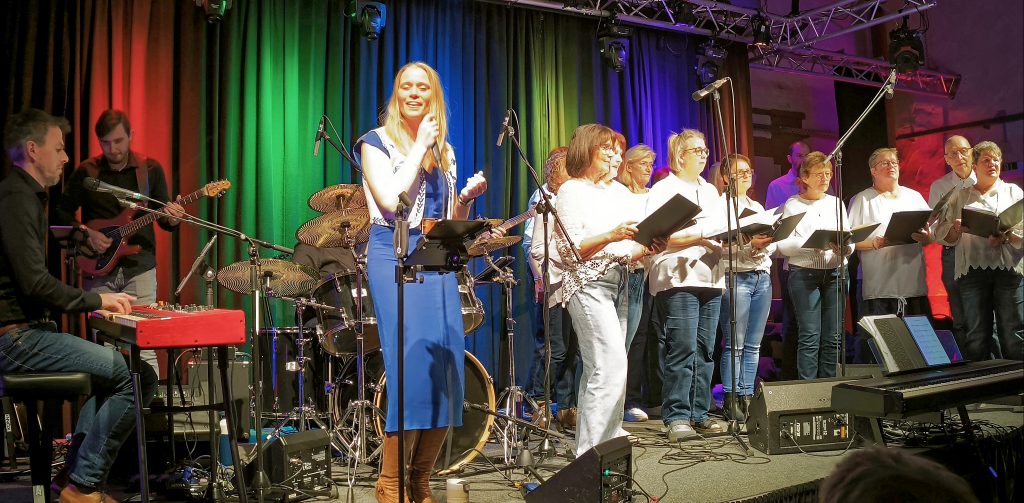 Eine blonde Sängerin mit langen glatten Haaren steht mit blauem Kleid auf der Bühne, das Mikrofon in der rechten Hand. Links hinter ihr ein Keyboarder und ein Gitarrist. Rechts von ihr ein in weiß gekleideter Chor (ca. 10-12 Personen). Der Vorhang hinter der Bühne ist mit breiten senkrechten Streifen roten, grünen, blauen und gelben Farben beleuchtet.