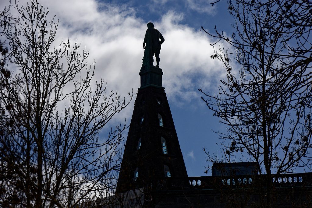 Eine überlebensgroße Statue auf einem spitz zulaufenden Sockel, der wiederum auf einem Gebäude (hier nicht zu sehen) steht. Dunkel im Gegenlicht. Blauer Himmel, weiße Wolken. Links und rechts ein Baum, noch ohne Blätter.