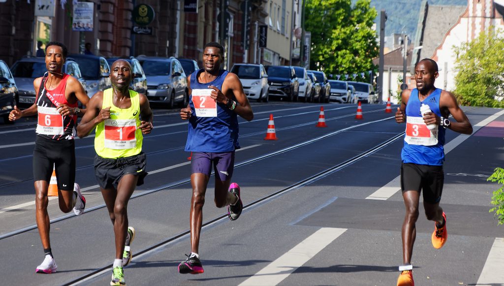 Vier Läufer in einer Reihe bei einem Stadtmarathon auf der Straße. Sie tragen von links nach rechts die Nummern 6, 1, 2 und 3.