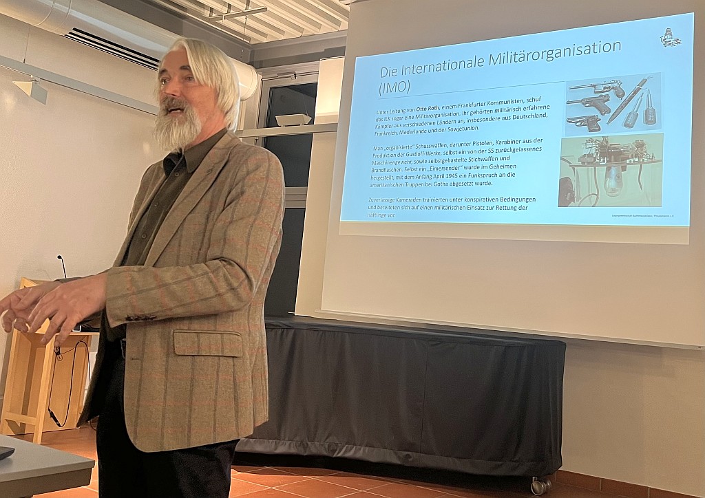 Ein älterer Mann mit langem grauen Bart und halblangen grauen Haaren, hellbraunes Jacket, steht an einem Pult und schaut nach links zum nicht sichtbaren Publikum. Rechts im Hintergrund eine Präsenationsfolie: "Die internationale Militärorganisation (IMO)"