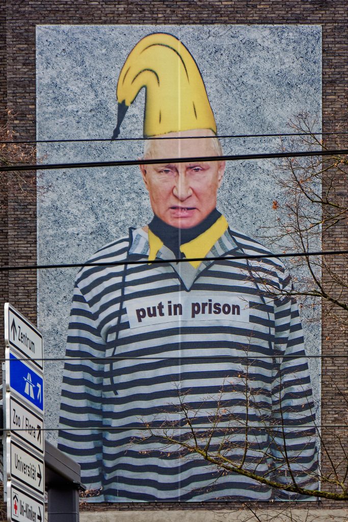 Putin in Gefängniskleidung = schwarz-weiß quergestreiftes Hemd mit einer Banane auf dem Kopf vor einem grauen Hintergrund an einer verklinkerten braunroten Hauswand. Im Vordergrund drei Oberleitungen. Rechts ragen Äste ins Bild. Links Straßenschilder, die von oben nach unten folgende Richtungen anzeigen:

Zentrum (geradeaus)
Autobahn (halbrechts)
Zoo / Flora ( " )
Universität (rechts)
Uni-Kliniken ( " )
