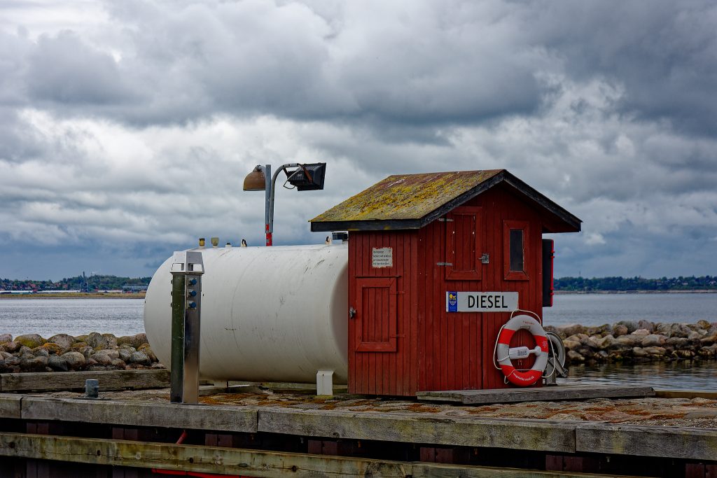 Ein rotes Holzhäuschen steht auf einem Anlieger in einem Yachthafen. Auf dem Holzhäuschen ein weißes Schild mit schwarzer Schrift Diesel. Darunter ein rot-weißer Rettungsring. Hinter dem Häuschen ein großer weißer Tank und eine Wasserfläche. Der Himmel ist mit dunklen Wolken verhangen.