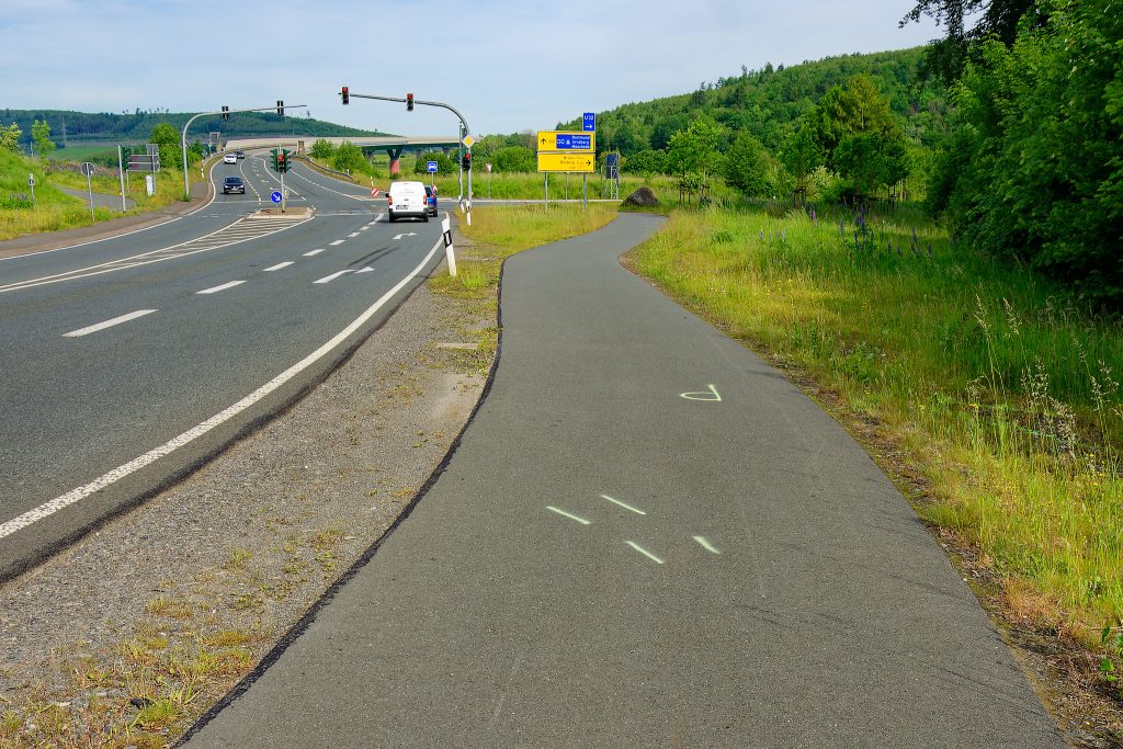 Ein breiter Radweg verläuft rechts neben einer Starße die auf eine Ampelkreuzung zuläuft. Auf dem Radweg eine mutmaßliche Unfallzeichnung: ein P und je zwei parallele Striche diagonal zum Weg.