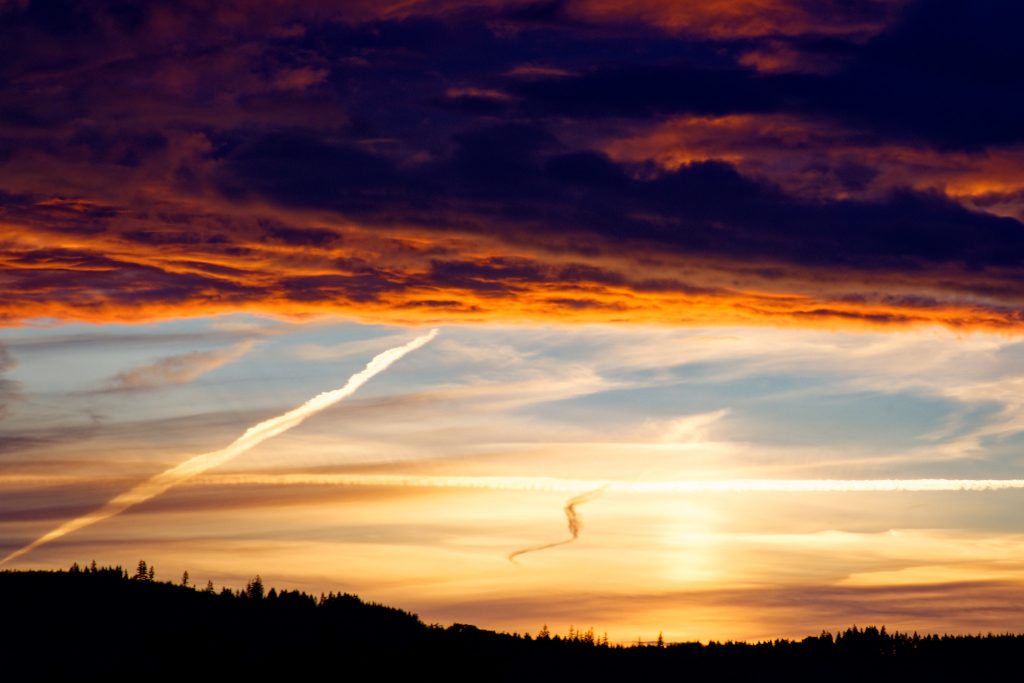 Ein dreigeteiltes Bild: unten die schwarze Hügelkette, darüber der blaugelbe Abendhimmel von leichten Wolken und Kondensstreifen durchzogen. Zum Abschluss oben eine bedrohlich wirkende dunkelrote Wolkendecke.