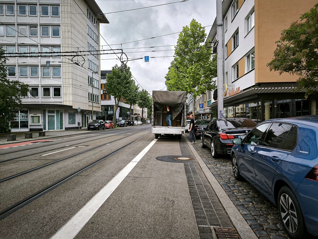 Eine städtische Straße mit Radschutzstreifen auf dem ein Liverwagen steht, der gerade entladen wird. Rechts eine Reihe von parkenden Autos, links Straßen- und Tram-Verkehr. Stromleitungen für die Trams über der Straße. Schienen in die Straße eingelassen. Neben der Straße hohe Häuser im 60er/70er- Jahre Stil.