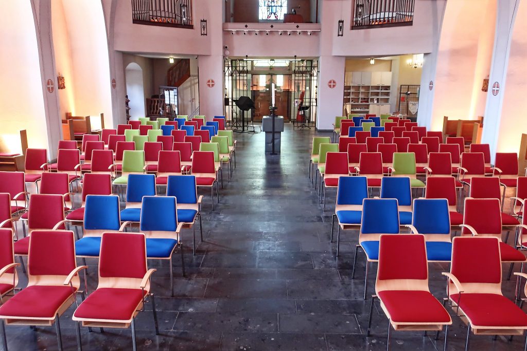 Blick in den Innenraum einer Kirche Richtung Altar. Links und rechts Stuhlreihen mit rot, grün und blau gepolstertem Gestühl. Der Boden ist schwarz, die Wände sind weiß.