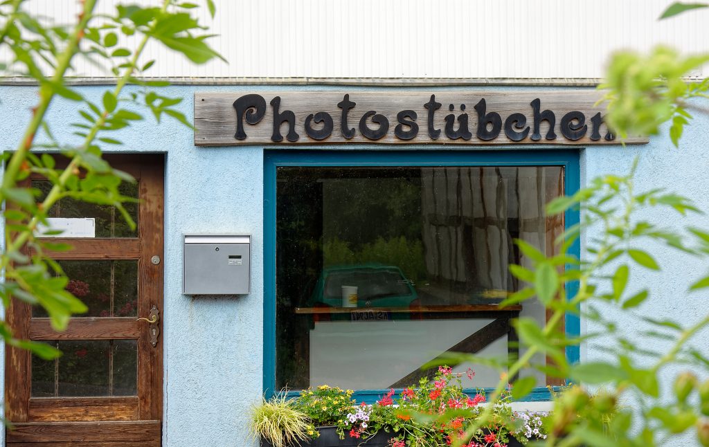 Die Frontseite eines Ladenlokals. Links eine brauen, hölzerne Eingangstür, rechts das Fenster, blauer Rahmen, mit buntem Blumenkasten unten. Dazwischen ein silberner Briefkasten. Über dem Fenster ein großes hölzernes Schild mit der dunklen Inschrift "Photostübchen". 

Im Vordergrund als Rahmen unscharf Zweige mit hellgrünen Blättern.